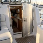 A cabin entry boat door on a cruising motor yacht, half open on its barrel hinges with a stainless slam latch, photographed in morning light at a marina.