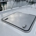 A walkable boat hatch set flush into a fiberglass deck after overnight rain, its aluminum lid closed on a continuous rubber gasket with stainless latches on top.