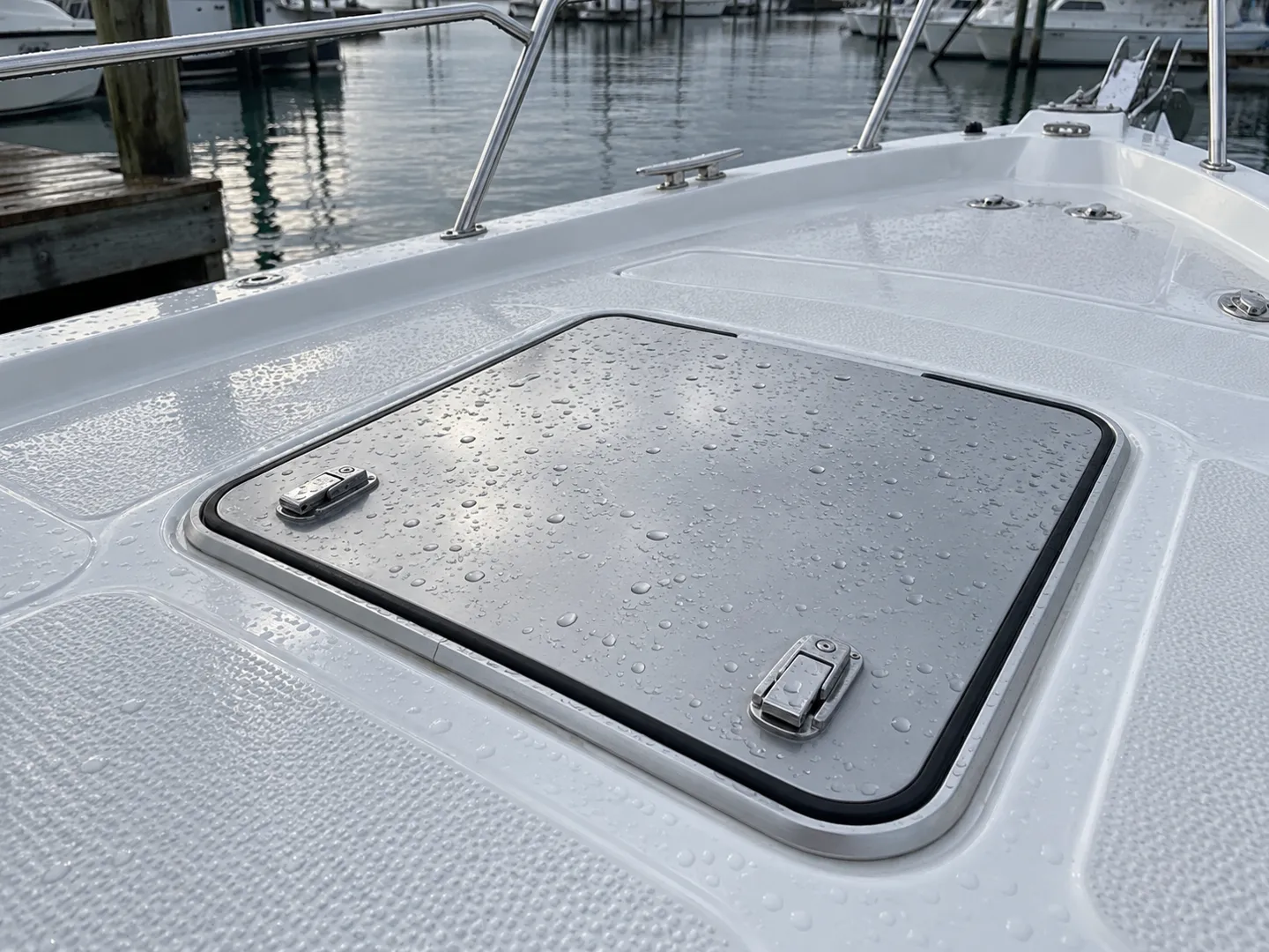 A walkable boat hatch set flush into a fiberglass deck after overnight rain, its aluminum lid closed on a continuous rubber gasket with stainless latches on top.